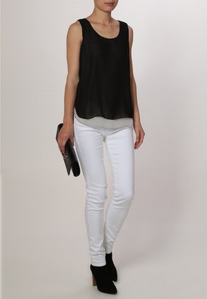 Schwarzes ärmelloses Top mit fließendem Design, über einer weißen Basis getragen, kombiniert mit weißen Skinny Jeans und schwarzen Knöchelstiefeln.