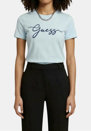 Persona che indossa una maglietta azzurro chiaro con "Guess" scritto in corsivo e pantaloni neri a vita alta, accessoriata con una collana a catena d’argento.