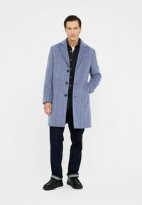 Cappotto in tweed blu con rever a punta, bottoni a un solo petto e tasche laterali, abbinato a jeans scuri e scarpe nere.