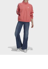 Pink fleece pullover with a high collar and white stripes, paired with flared blue jeans and white shoes with black accents.