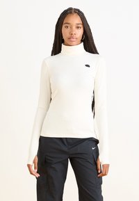 Top de gola alta em creme com textura de riscas, apresentando mangas longas e um pequeno logótipo preto no peito, combinado com calças cargo pretas.