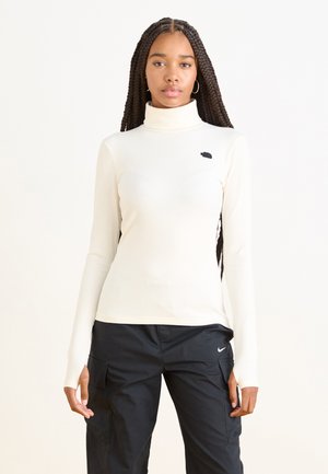 Kremowy sweter z golfem o prążkowanej fakturze, z długimi rękawami i małym czarnym logo na piersi, zestawiony z czarnymi spodniami cargo.