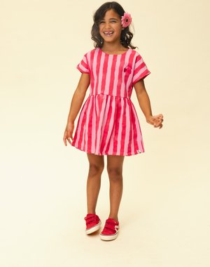 Lachend meisje met donker krullend haar en een roze bloem, gekleed in een rood- en roze gestreepte jurk en rode sneakers, staand op een crèmekleurige achtergrond.