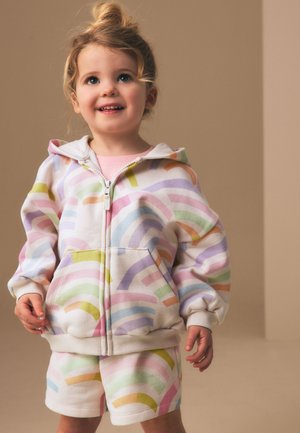 Mladá dívka se světlými vlasy svázanými do drdolu, oblečená v pastelově duhové zipové mikině a odpovídajících šortkách, usmívající se na béžovém pozadí.