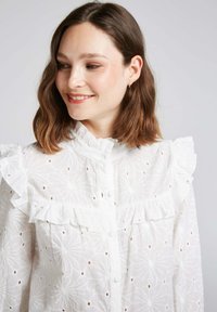Blouse blanche brodée avec des accents froncés, présentant un col haut et une fermeture à boutons. Motif floral avec de petites découpes pour ajouter de la texture.