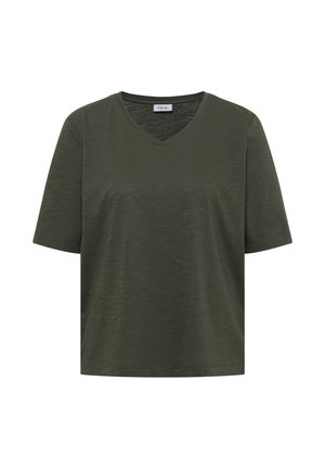 Kvinde smiler, står i et studie, iført en løs mørkegrøn V-hals T-shirt, blå jeans og beige sneakers med grønne detaljer.
