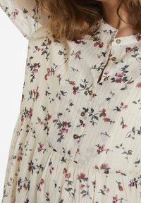 Detailní záběr na krémové šaty s tenkými zlatými pruhy a malými růžovými a černými květinovými vzory, které mají zlaté knoflíky po celé přední straně.
