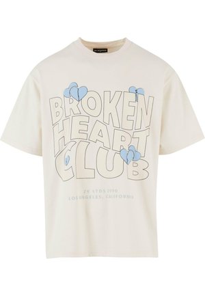 T-shirt en coton couleur crème avec un col rond, arborant en grand texte noir "BROKEN HEART CLUB" et des graphiques de cœurs bleus sur le devant.