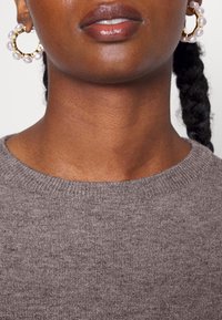 Grauer Strickpullover mit rundem Ausschnitt. Große Creolen mit einem goldfarbenen Metallrahmen und perlenähnlichen Akzenten. Glatte Hautstruktur sichtbar.