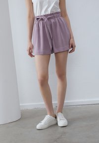 Shorts lavande avec une finition texturée, taille élastique et détail de nœud à l'avant. Associés à des baskets blanches sur un sol en béton.