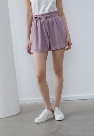 Lavender shorts with a textured finish, elastic waistband, and front tie detail. Paired with white sneakers on a concrete floor.