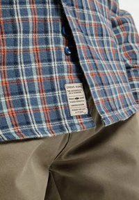 Koszula w kratę w kolorach niebieskim, pomarańczowym i białym, z metką, zapięciem na guziki i miękką fakturą; noszona nad beżowymi spodniami.