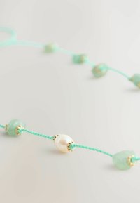 Ett pärlhalsband med ljusgröna stenar, en central vit pärla och guldaccenter på en turkosa snodd.