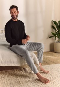 Camicia nera a maniche lunghe e pantaloni da pigiama grigio chiaro su un uomo seduto su un letto, con una parete beige e una pianta verde sullo sfondo.