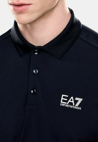 Camisa polo navy con un cuello texturizado, tres botones y un logo de EA7 Emporio Armani en blanco. Tejido suave con un diseño clásico.
