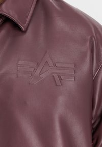 Detail obrazu fialovej koženej bundy s embosovaným geometrickým logom, ktoré zobrazuje trojuholník a horizontálne pruhy na hrudi.