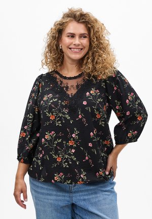 Woman with curly blonde hair smiling, wearing a black floral blouse with lace detail and blue jeans, standing with one hand on hip.