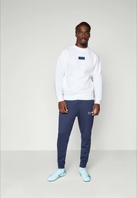 Sweat-shirt blanc avec un logo en patch bleu, associé à un pantalon de jogging marine avec un swoosh Nike blanc et des chaussures de sport bleu clair.