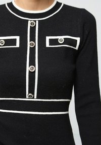 Robe en maille noire avec des accents blancs, col rond, fermeture par bouton et poches décoratives. Tissu texturé avec une forme structurée.