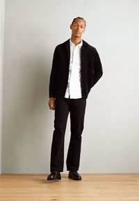 Veste en velours côtelé noire, chemise blanche à boutons, pantalon noir et chaussures noires brillantes. Textures lisses, design classique et coupe ajustée.