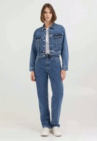 Casaco de denim e jeans de cintura alta em azul. O casaco possui dois bolsos no peito e um design cropped. A modelo usa ténis brancos.