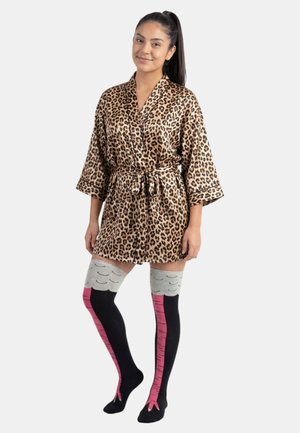 Vrouw draagt een badjas met luipaardprint en zwarte kousen tot boven de knie met een ontwerp van roze flamingopoten, staand tegen een effen achtergrond.