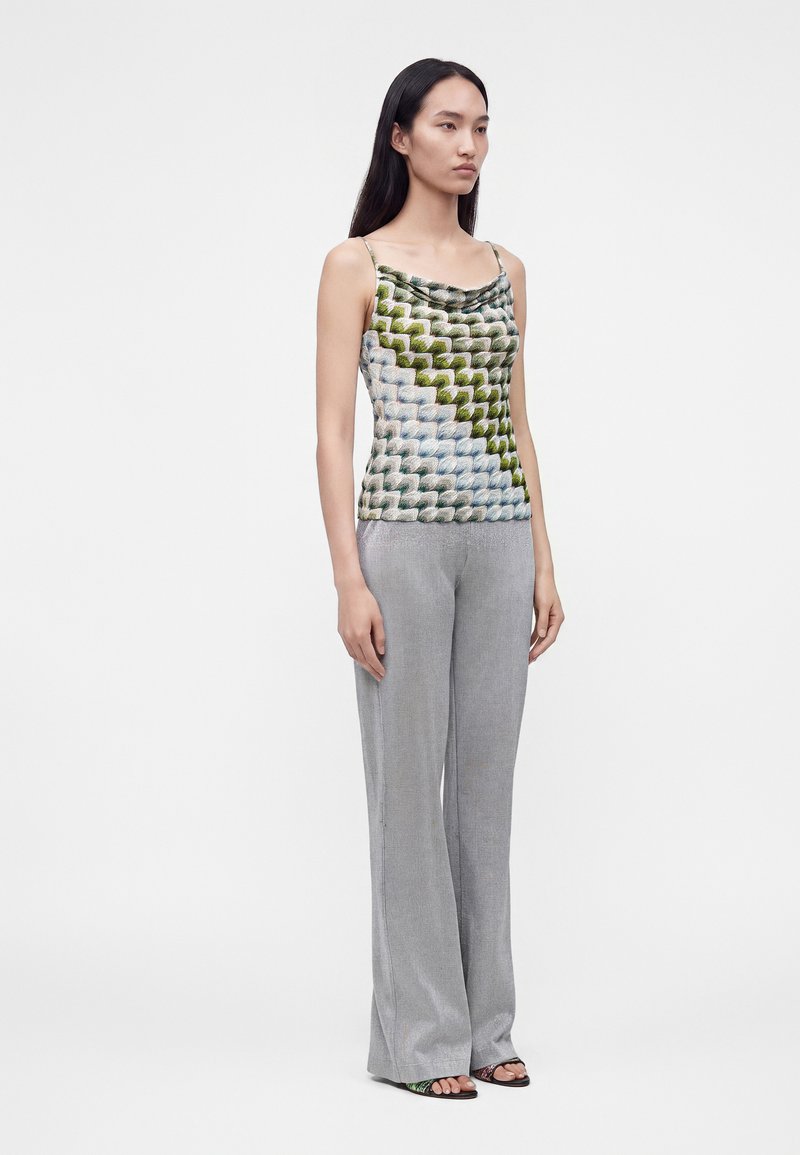 Femme debout portant un top sans manches à motif avec des bretelles fines et un pantalon large gris clair avec des sandales ouvertes.