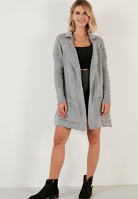 Cardigan gris con canalé, de frente abierto, con dos bolsillos y tela texturizada. Se lleva sobre un top corto negro y una falda de cuero, combinado con botines negros.