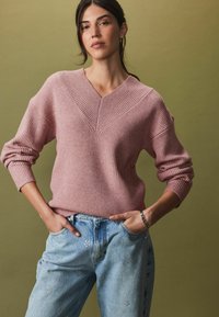 Vaaleanpunainen ribbikuvioitu neulepusero, jossa on V-pääntie, yhdistettynä vaaleansinisiin farkkuihin, joissa on koristeellisia yksityiskohtia. Taustana on tasainen vihreä.