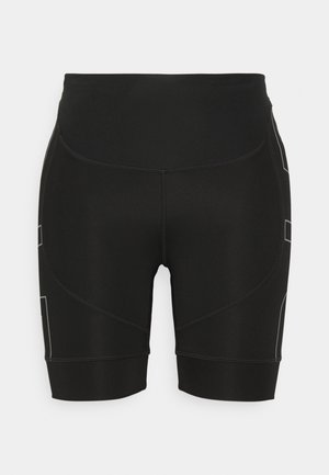 Femme en brassière de sport noire et short tenant une bouteille d'eau rose, portant des chaussures de course bleues et violettes, marchant devant un fond uni.