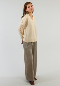 Maglione lavorato a maglia crema con scollo a V e dettagli in pizzo decorativo sulle maniche. Abbinato a pantaloni marroni larghi e texture.