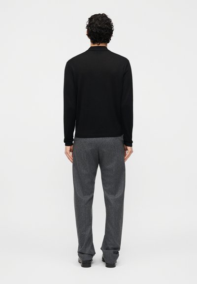 Άνδρας που στέκεται πλάτη στην κάμερα, φοράει μαύρο μακρυμάνικο πουκάμισο, γκρι παντελόνι και μαύρα παπούτσια, πάνω σε απλό λευκό φόντο.