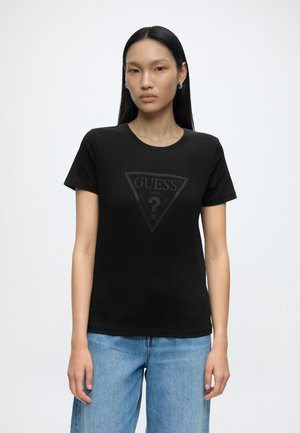 Kvinde med langt sort hår iført sort Guess t-shirt og blå jeans, står foran en ensfarvet hvid baggrund.