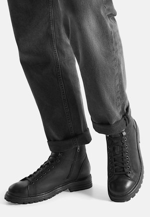 Czarne skórzane buty na gumowej podeszwie, z przodem na sznurowadła, bocznym zamkiem błyskawicznym i zaokrąglonym noskiem, w parze z podwiniętymi szarymi spodniami.