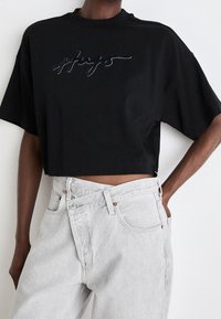 Černé zkrácené tričko s kulatým výstřihem a krátkými rukávy, na kterém je nápis "hugs" s lesklou aplikací. V kombinaci se světle šedými džíny s vysokým pasem.