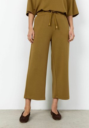 Asmuo dėvi laisvus alyvuogių žalios spalvos platėjančius trumpus kelnes su raišteliu juosmens srityje ir tamsiai rudos spalvos lygiais batais, stovintis prie baltos sienos.