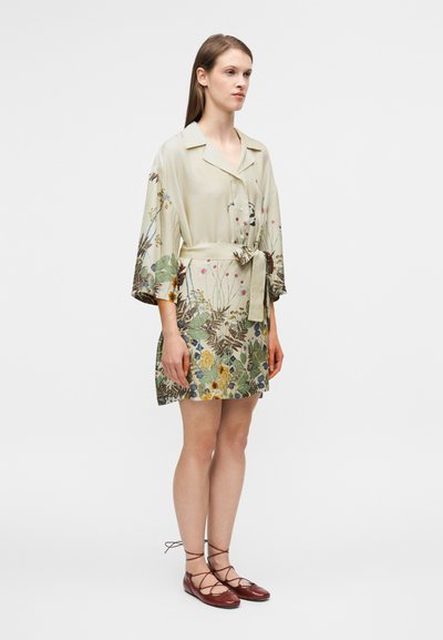 Robe de couleur claire à imprimé floral avec un col châle, une taille ceinturée et des manches évasées ; associée à des ballerines marron à lacets.