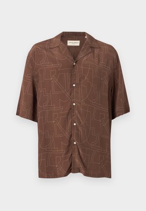 Άνδρας όρθιος, φοράει καφέ πουκάμισο με κοντά μανίκια και μοτίβο, μπεζ παντελόνι με φαρδιά μπατζάκια, μαύρα σανδάλια και λευκό φανελάκι, κοιτάζει πλευρικά.