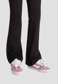 Sneaker in suede rosa con lacci bianchi e suole in gomma. Presentano una striscia laterale bianca e dettagli neri a contrasto. Indossati con pantaloni neri.