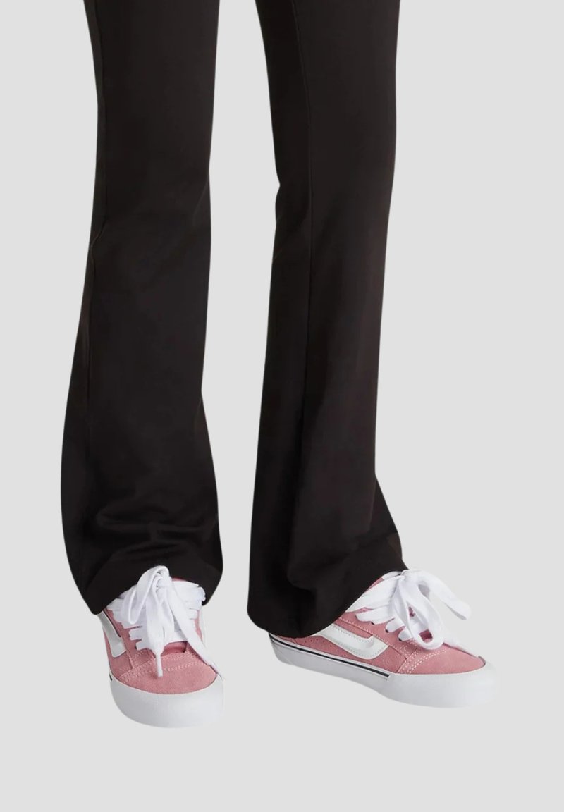 Sneaker in suede rosa con lacci bianchi e suole in gomma. Presentano una striscia laterale bianca e dettagli neri a contrasto. Indossati con pantaloni neri.