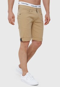 Beige denim shorts med opkørte manchetter, kontrasterende mørkeblå striber og en Calvin Klein logo elastik. Båret med hvide sneakers.
