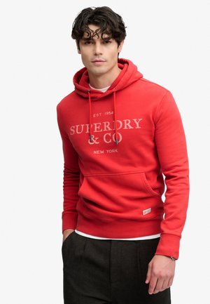 Ung mand med mørkt krøllet hår iført en rød hættetrøje med teksten Superdry & Co, hænderne i lommerne, på en ensfarvet hvid baggrund.