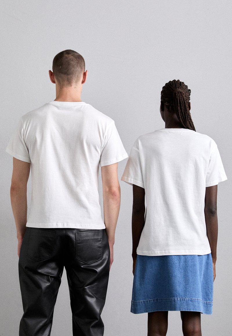 Two individuals wearing white cotton t-shirts, standing back-to-back. One person wears black leather pants, the other a denim skirt.