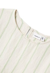 Tessuto di colore crema con strisce verticali verde chiaro, scollatura rotonda e una piccola apertura sul retro. Etichetta visibile all'interno.