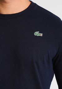 Camisa de manga larga azul marino de algodón, que presenta un pequeño logo de cocodrilo verde en el pecho izquierdo. Textura suave con diseño de cuello redondo.