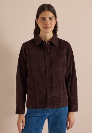 Femme aux longs cheveux foncés portant une veste en velours côtelé marron foncé et un jean bleu, debout devant un fond beige.