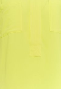 Helles gelbes Material mit zwei frontalen Taschen, einer vertikalen Knopfleiste und einer glatten Textur. Der Stoff wirkt leicht und leicht glänzend.