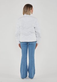 Giacca puffer bianca con motivo testurizzato, colletto alto e polsini a coste. Indossata con jeans blu a zampa d'elefante, che mostrano una silhouette aderente da dietro.