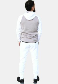 Sudadera gris y blanca con cremallera frontal, tela texturizada y un bolsillo canguro. Combinada con pantalones de chándal blancos y zapatillas negras.