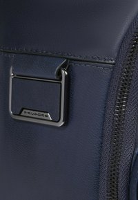 Marineblauwe leren tas met een gladde textuur, voorzien van een metalen gespsluiting en een contrasterende zwarte ritssluiting. Merkentag zichtbaar.